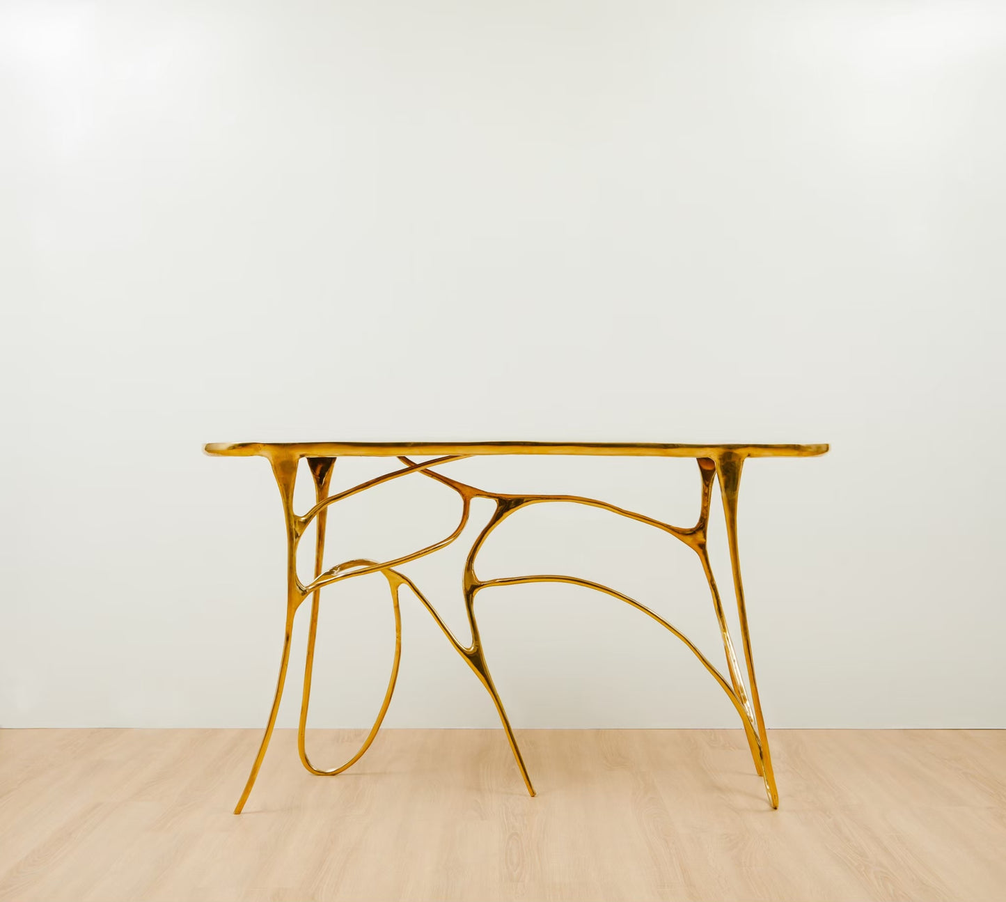 A modern sculptural console table with a polished gold metallic frame and a rectangular top, set against a minimalist white wall and light wood floor.