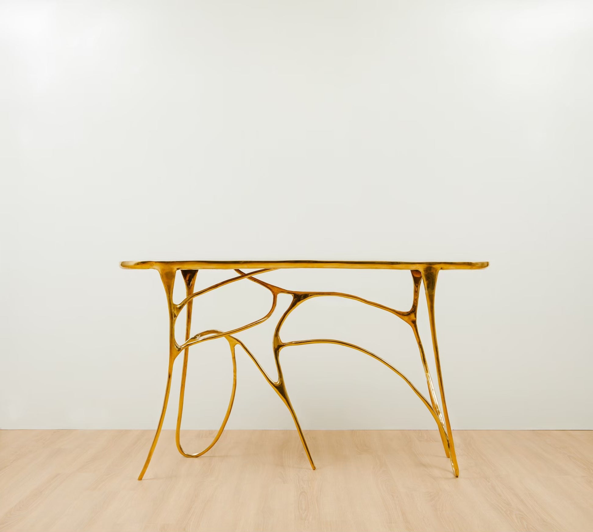 A modern sculptural console table with a polished gold metallic frame and a rectangular top, set against a minimalist white wall and light wood floor.