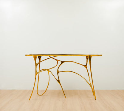 A modern sculptural console table with a polished gold metallic frame and a rectangular top, set against a minimalist white wall and light wood floor.