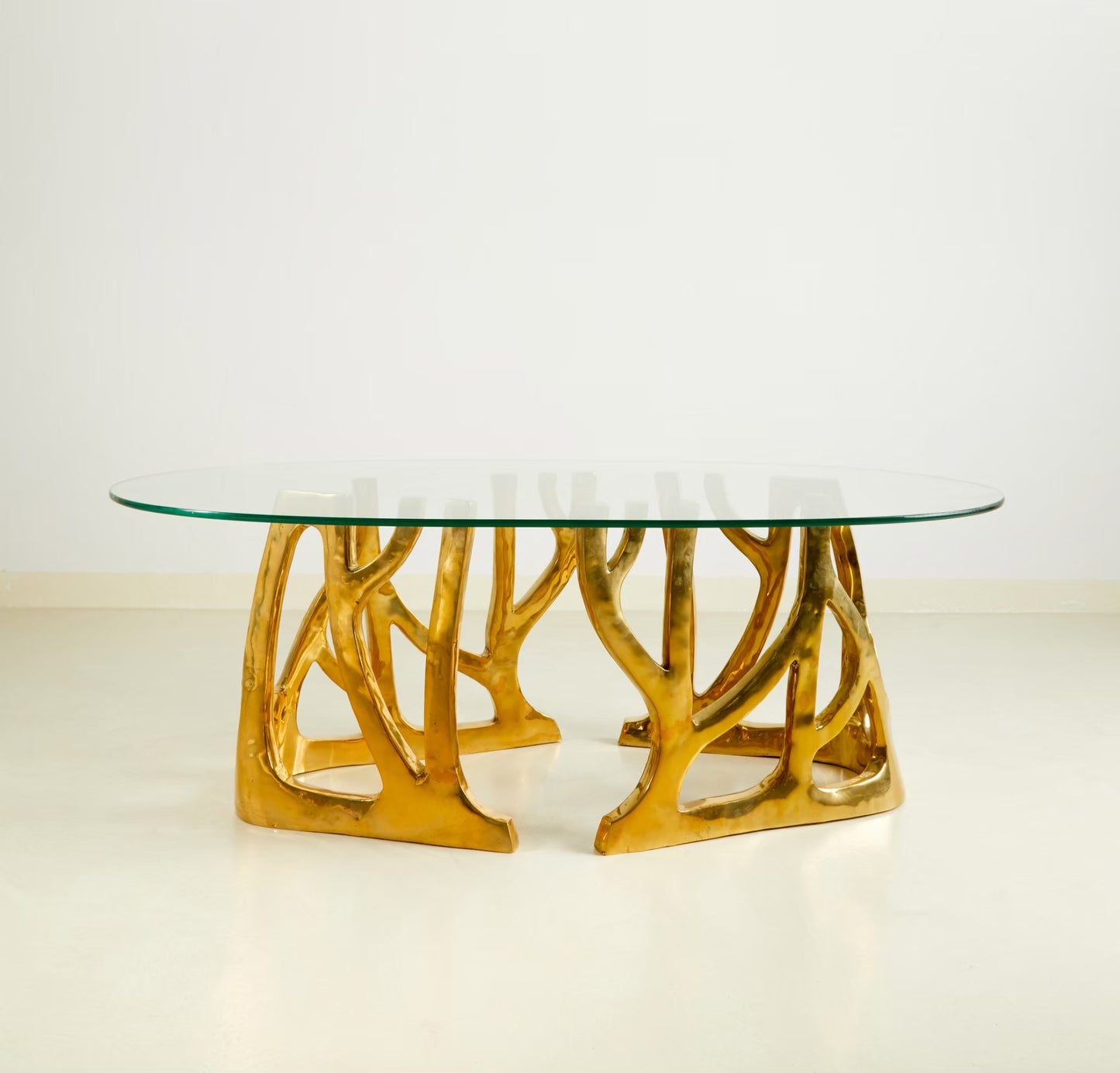 An oval coffee table with a clear glass top and a sculptural, branch-like metallic gold base, photographed against a clean white studio background.