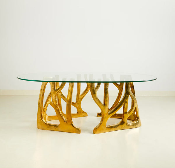 An oval coffee table with a clear glass top and a sculptural, branch-like metallic gold base, photographed against a clean white studio background.