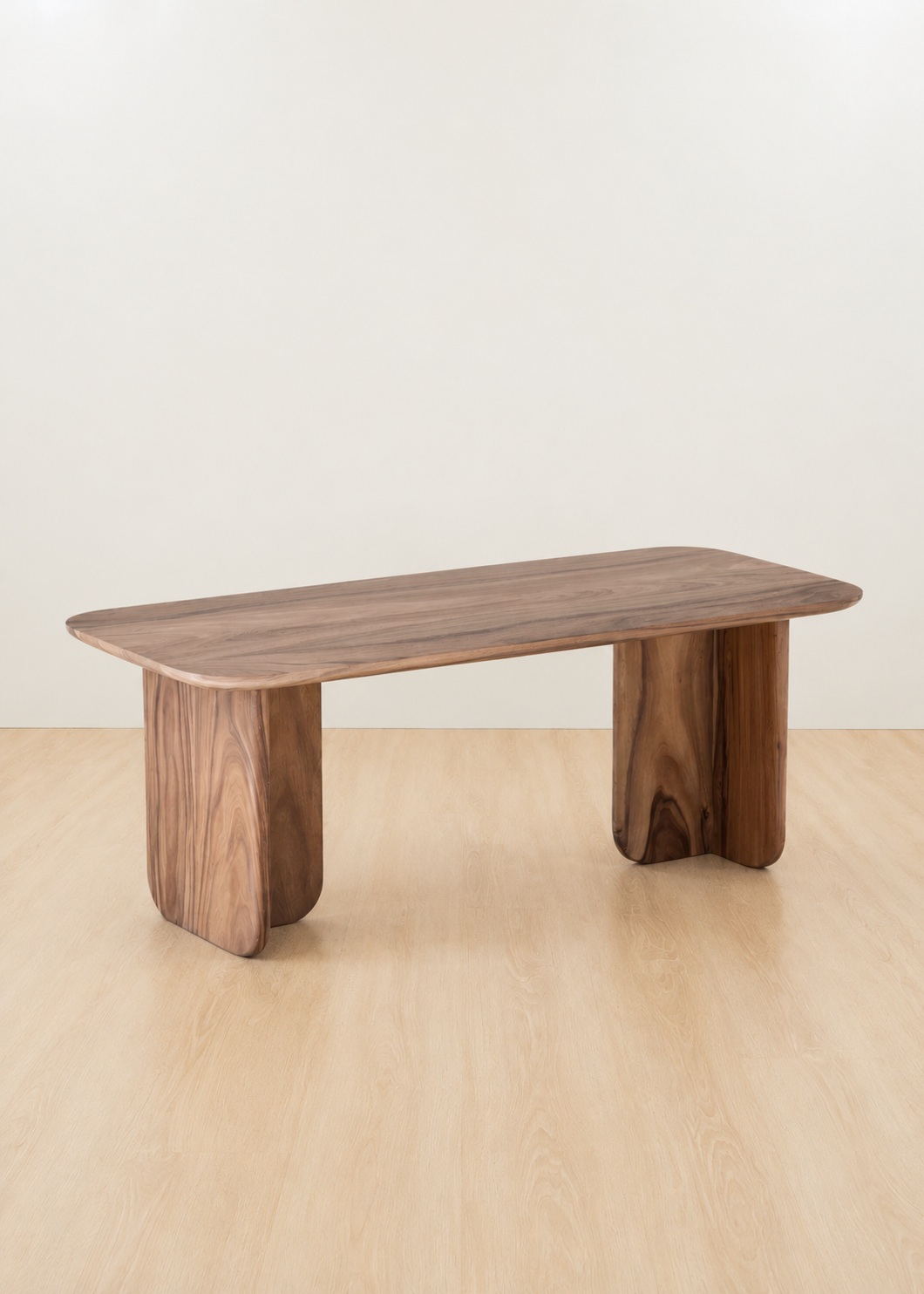 A modern wooden bench with a rounded rectangular top and two thick, blocky legs, shown on a light wood floor against a plain off-white wall.