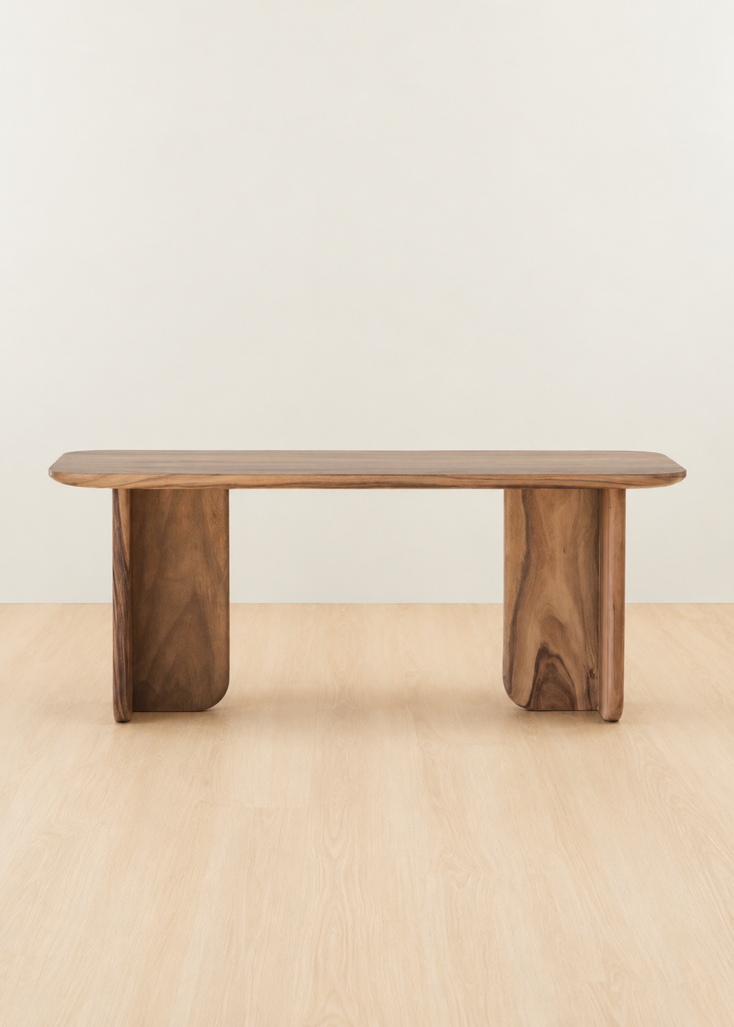 A minimalist wooden bench with a rounded rectangular top and two thick blocky legs, shown on a light wood floor against a plain off-white wall.