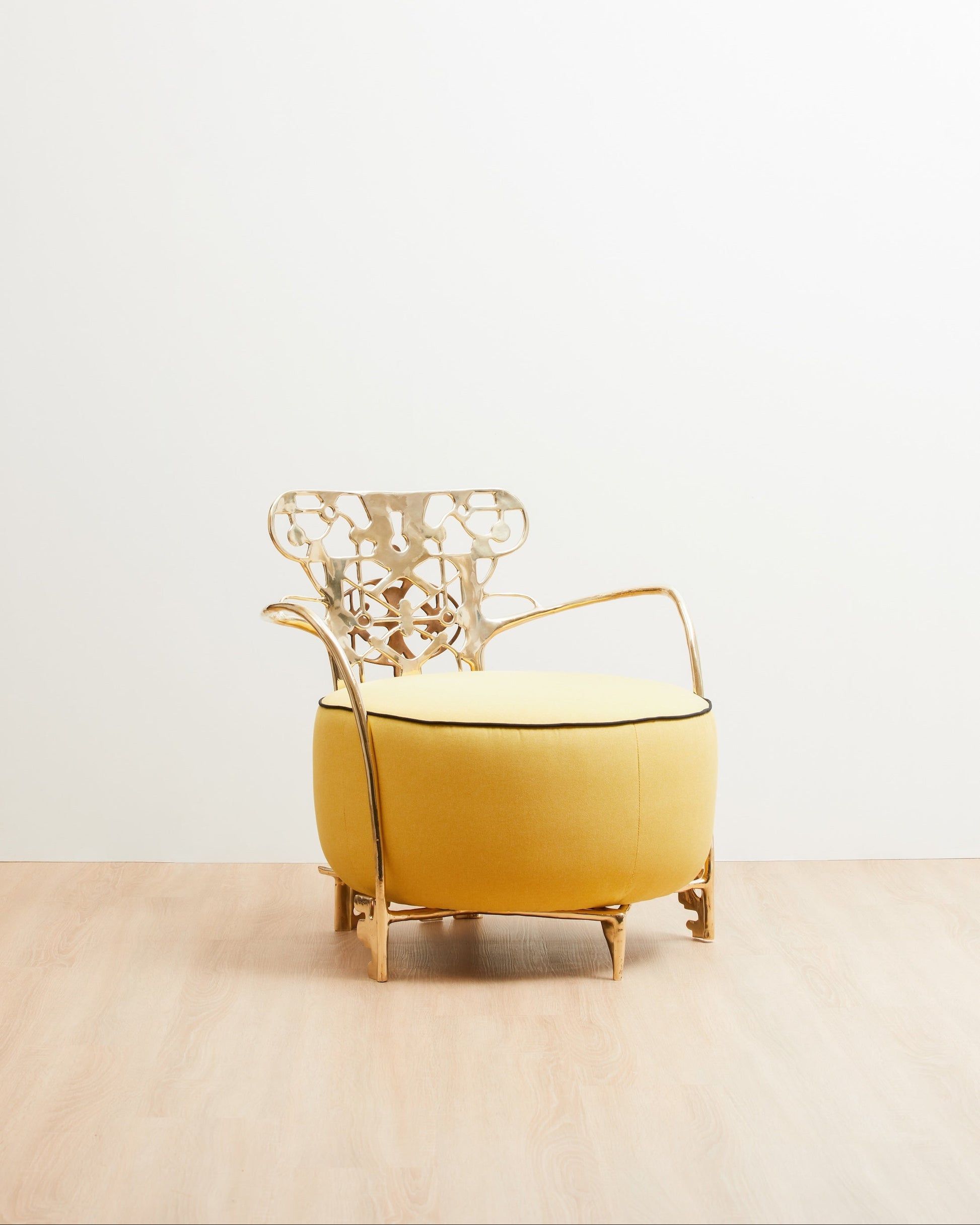 A modern accent chair with a round mustard yellow upholstered base and a sculptural gold metal frame, set against a minimalist white wall and light wood floor.