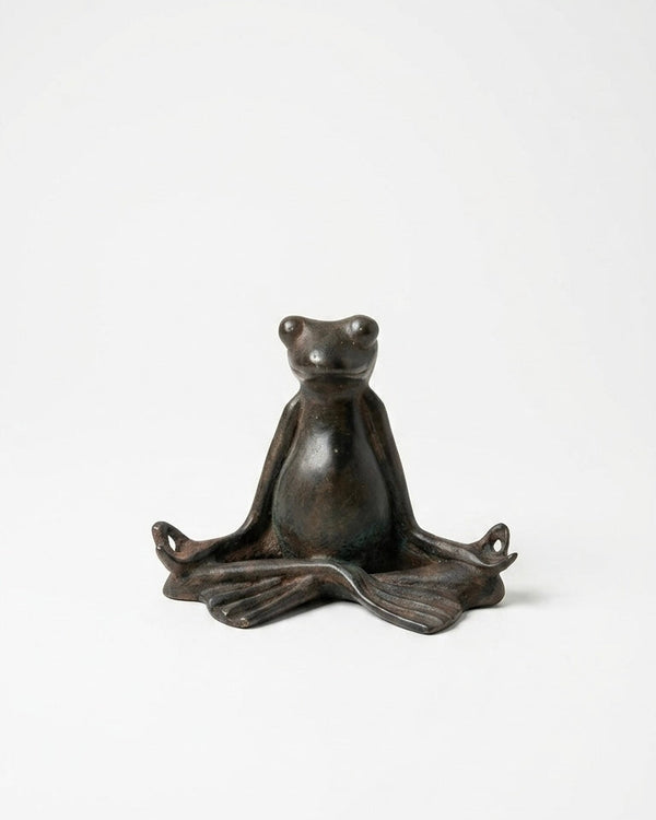 A small dark bronze frog figurine sitting in a meditative lotus pose, photographed against a seamless white studio background.