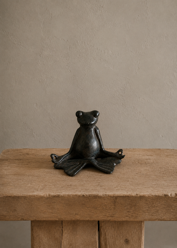 A small bronze frog figurine sitting cross-legged on a rustic wooden table against a textured beige wall.