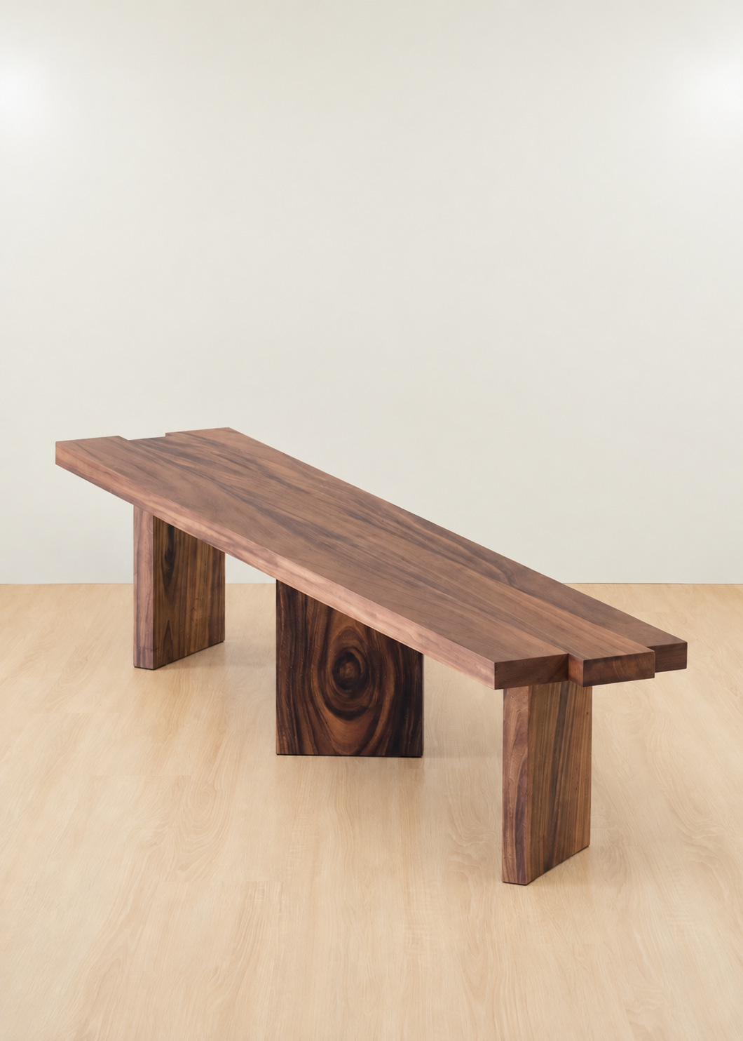 A rustic wooden bench with a live-edge top and block-style legs, shown in a minimalist studio setting with a light beige wall and pale wood floor.