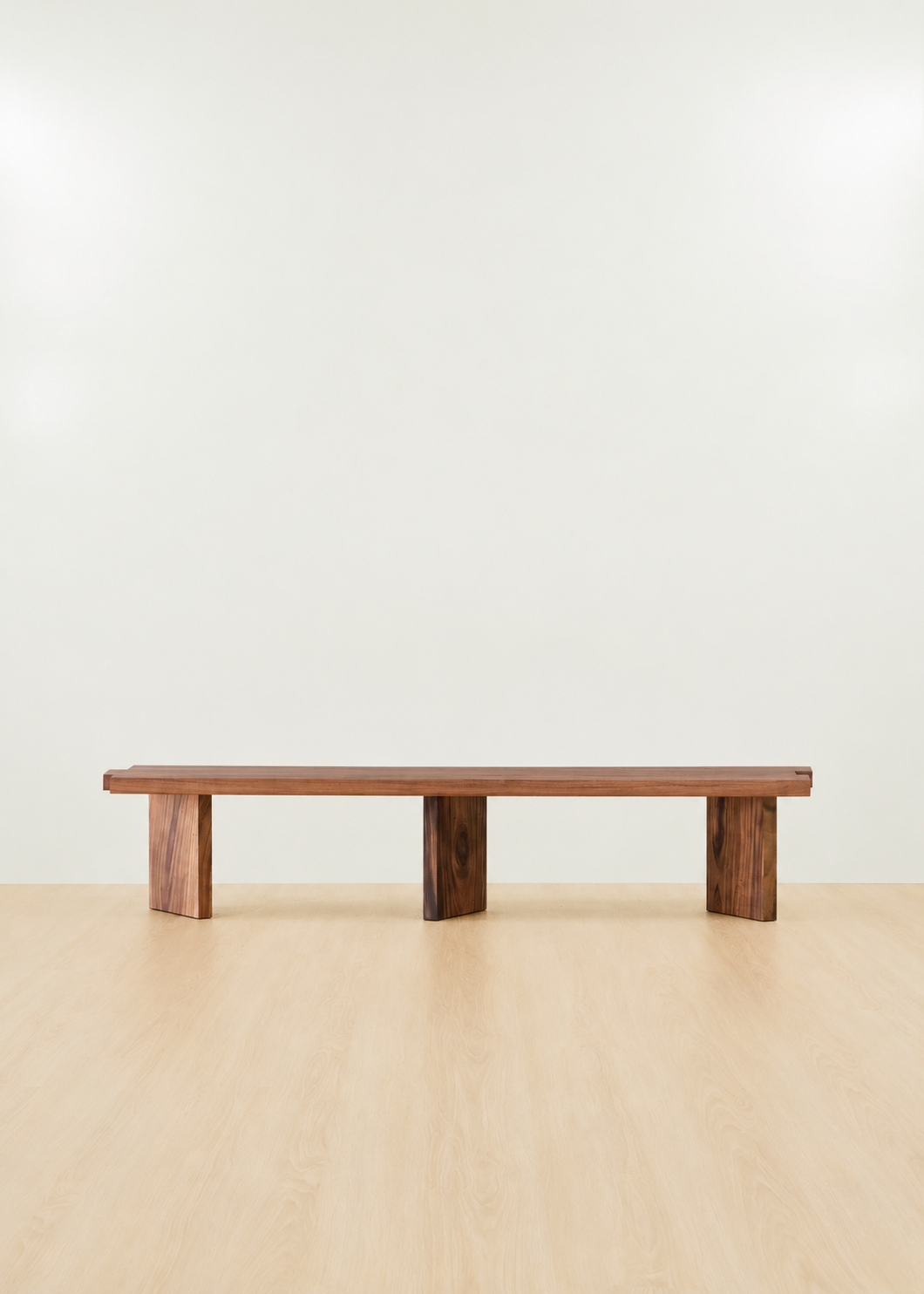 A minimalist wooden bench with a rectangular top and two block legs, shown on a light wood floor against a plain off-white wall.