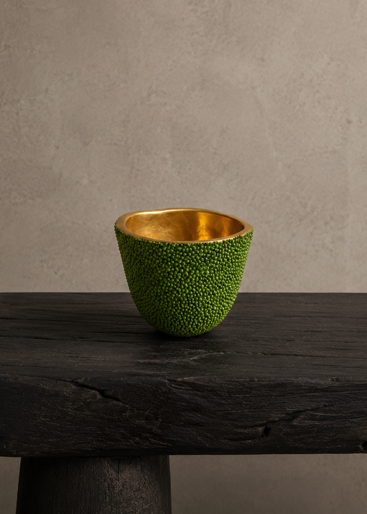 A small textured green cup with a shiny gold interior, displayed on a dark rustic wooden table against a neutral textured gray wall.
