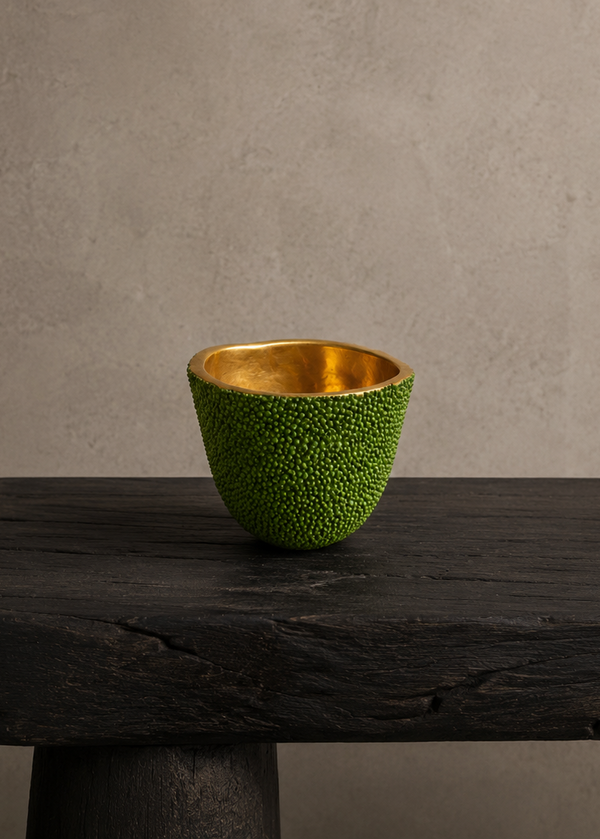A small textured green cup with a shiny gold interior, displayed on a dark rustic wooden table against a neutral textured gray wall.