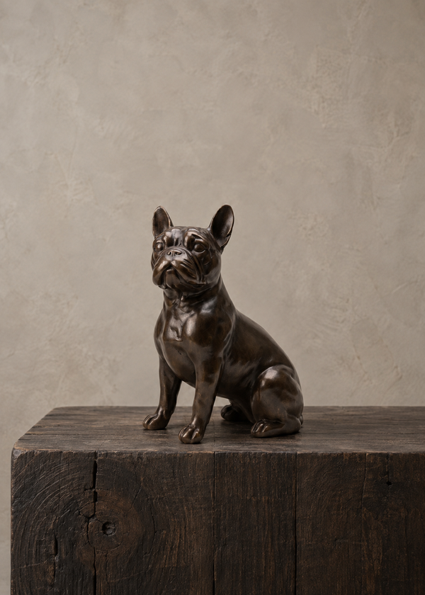 A dark bronze sculpture of a French Bulldog sitting on a rustic dark wood block, set against a textured beige plaster wall.