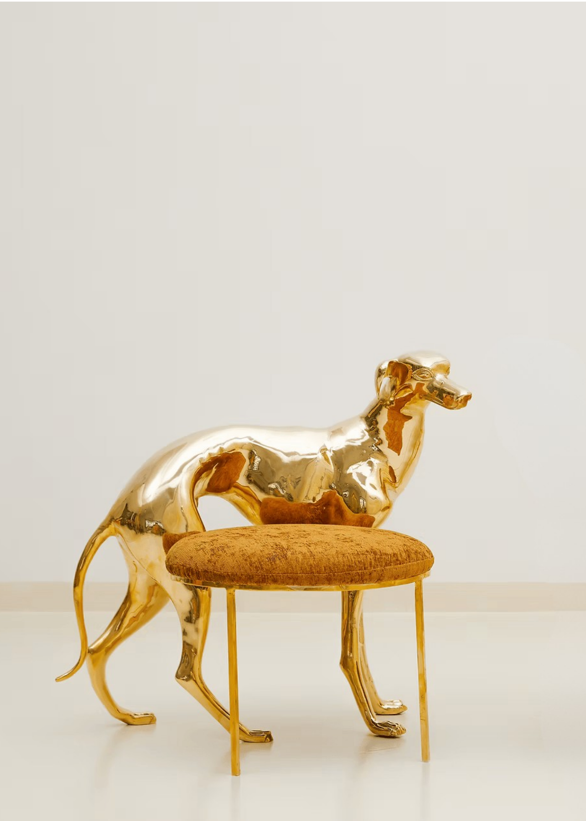 A polished brass dog sculpture posed behind a small round stool with a textured mustard-gold cushion and slim gold legs, set against a clean off-white studio background.