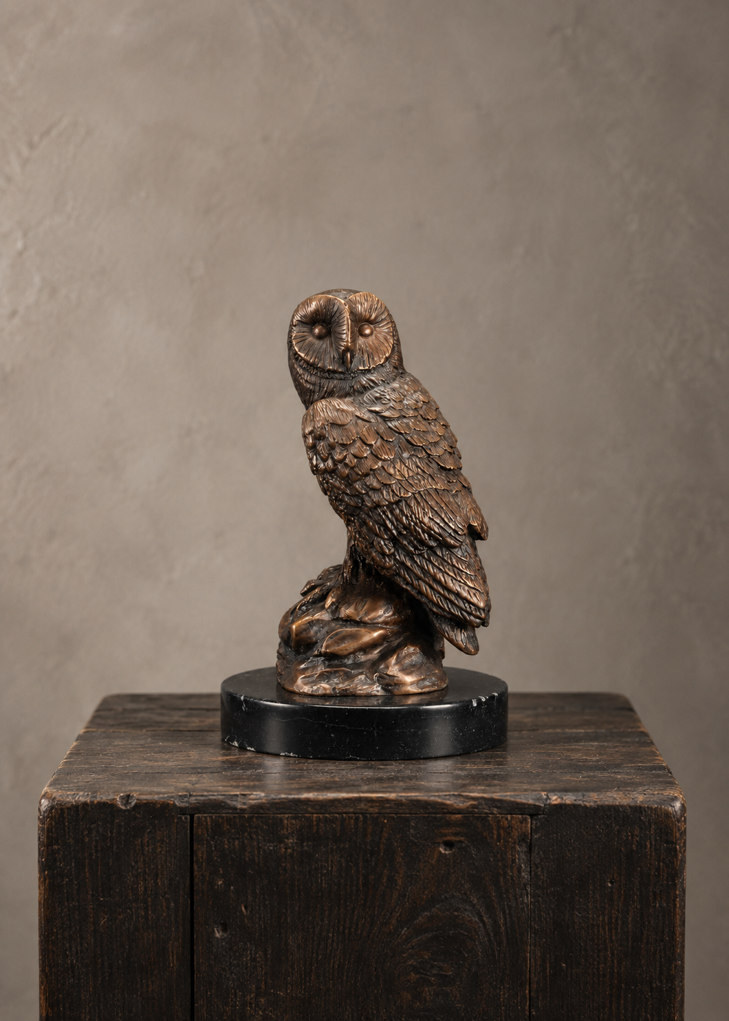A detailed bronze owl sculpture perched on a branch, mounted on a round black base and displayed on a rustic dark wooden pedestal against a textured gray background.