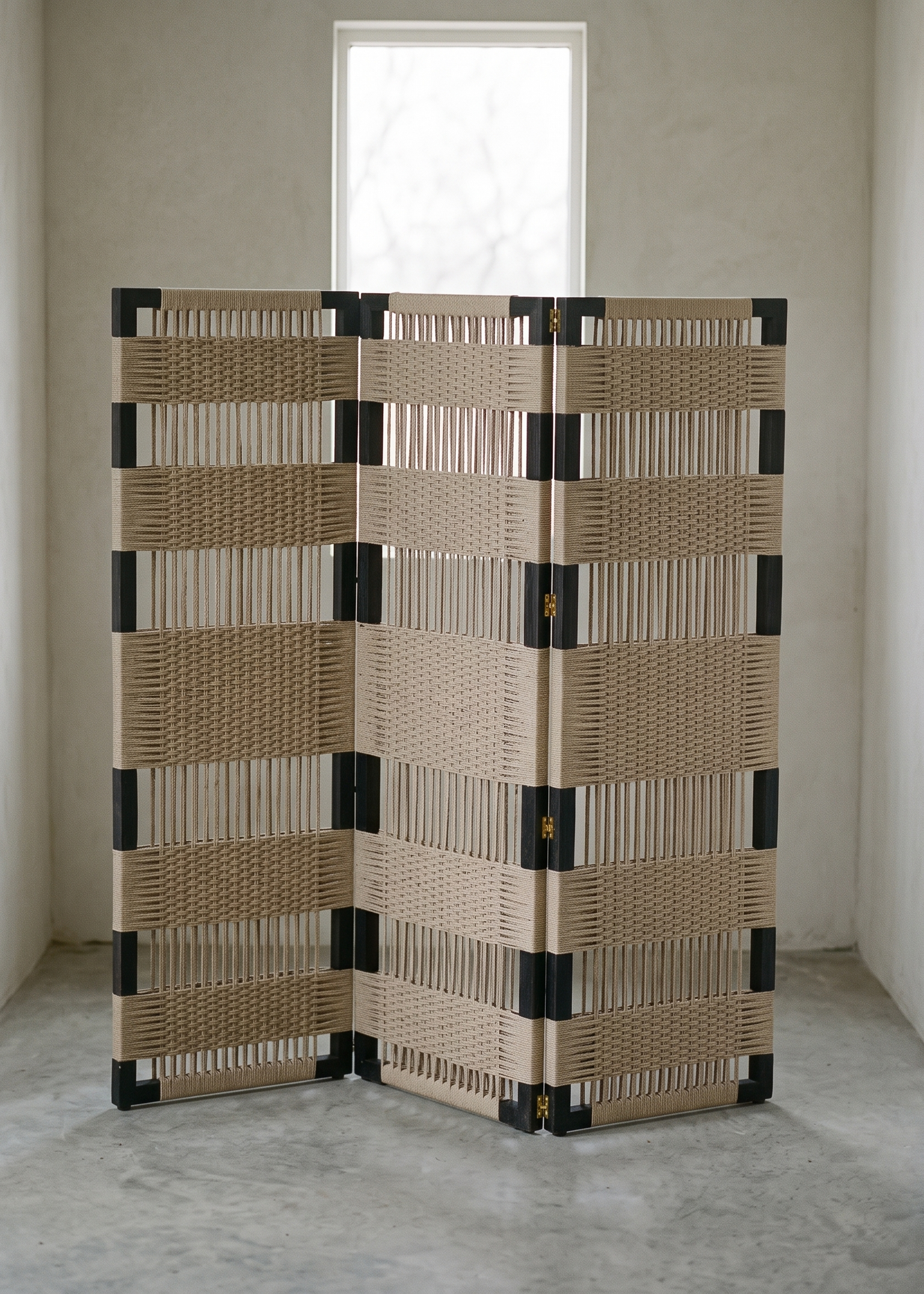 A folding screen with woven beige panels and black corner supports, standing in a minimalist room with concrete floors and plaster walls.