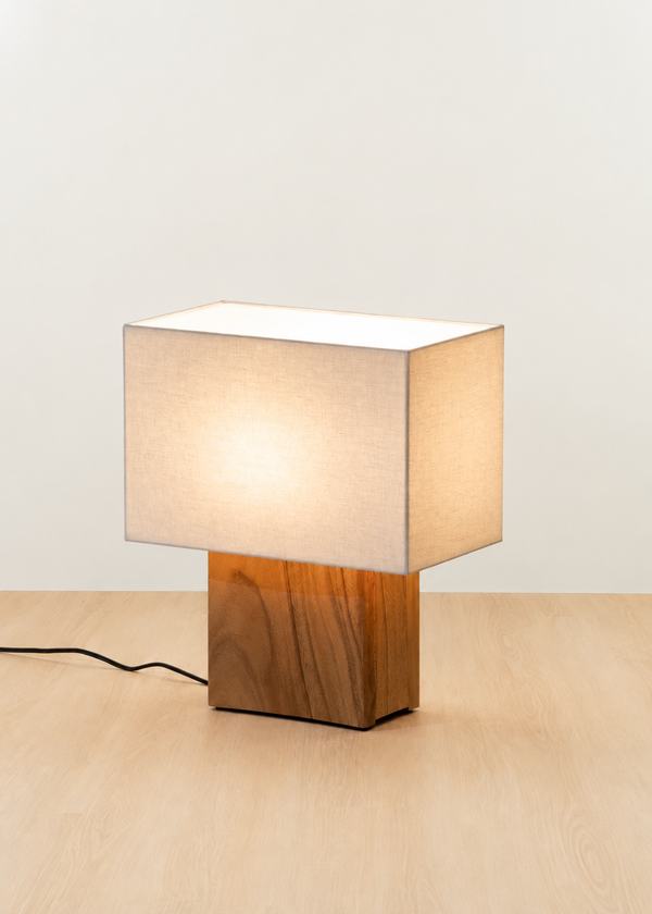 A modern table lamp with a square warm-white fabric shade and a rectangular wood-grain base, shown lit on a light wood surface against a plain off-white wall.