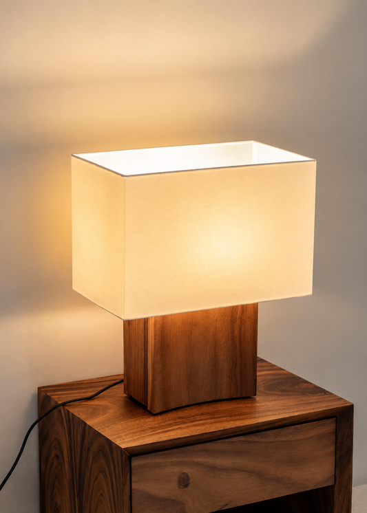 A modern table lamp with a square, warm-white illuminated fabric shade and a carved wooden base, shown resting on a matching wooden nightstand against a neutral wall.