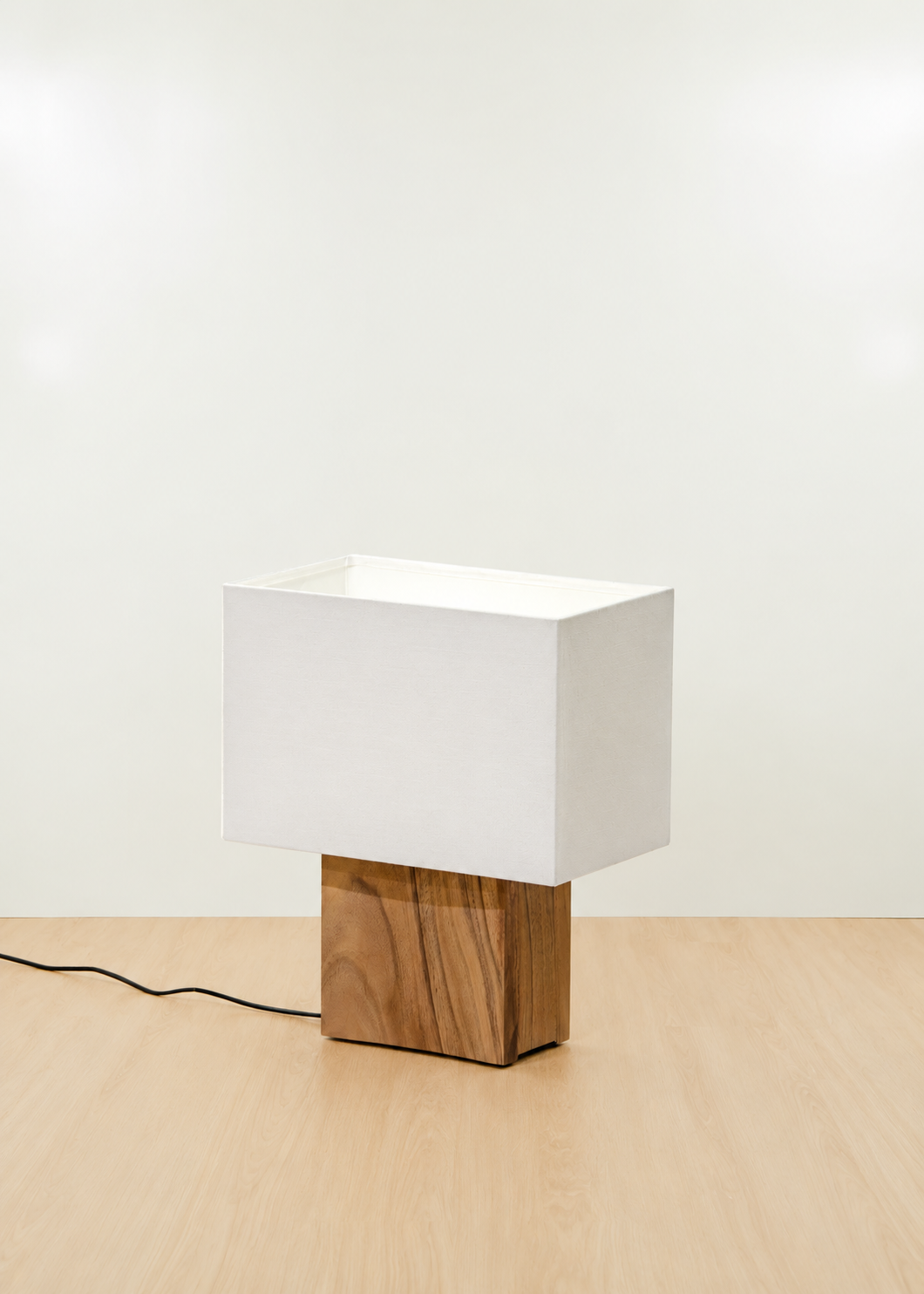 A modern table lamp with a white rectangular fabric shade and a square wooden base, set on a light wood floor against a plain white wall.