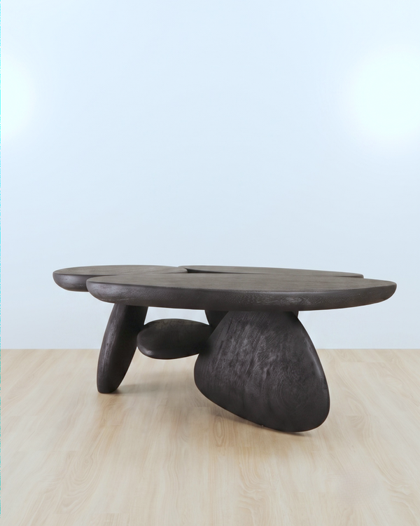 A dark carved wooden coffee table with an organic, organic-shaped top and three sculptural legs, set on a light wood floor against a pale blue background.