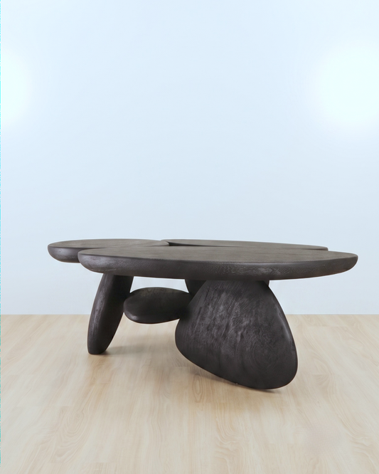 A dark carved wooden coffee table with an organic, organic-shaped top and three sculptural legs, set on a light wood floor against a pale blue background.