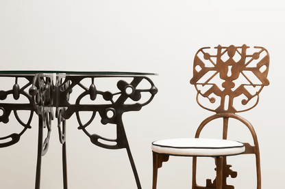 A close-up of a decorative metal dining table and matching chair with an intricate cutout backrest, set against a plain off-white background.