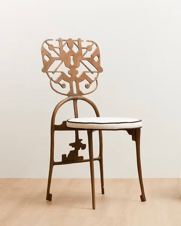 A modern brass dining chair with a sculptural cutout backrest, curved metal frame, and a white cushioned seat with a thin black edge, set against a minimalist beige wall and light wood floor.