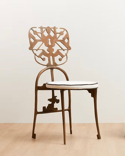 A modern brass dining chair with a sculptural cutout backrest, curved metal frame, and a white cushioned seat with a thin black edge, set against a minimalist beige wall and light wood floor.