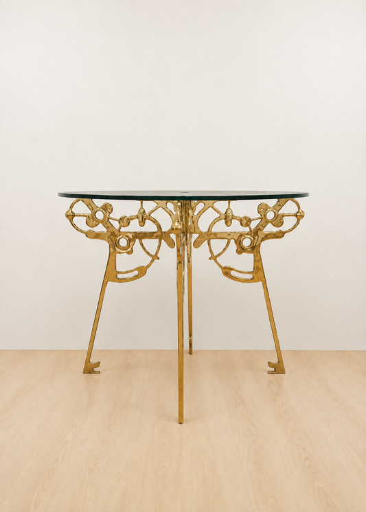 A round table with a clear glass top and an ornate gold brass base, set against a plain off-white wall and light wood floor.