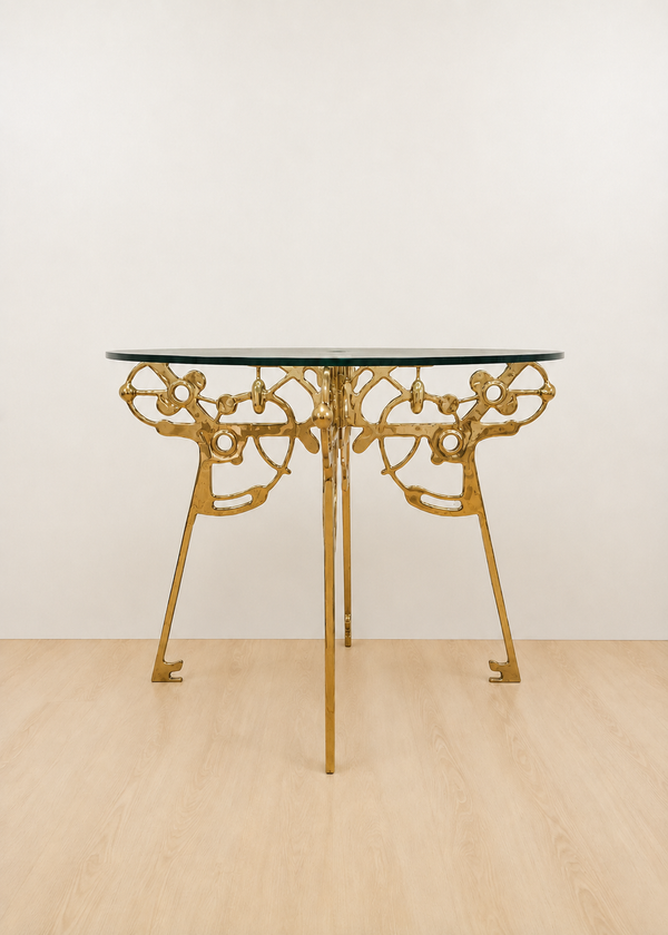 A round table with a clear glass top and an ornate gold brass base, set against a plain off-white wall and light wood floor.