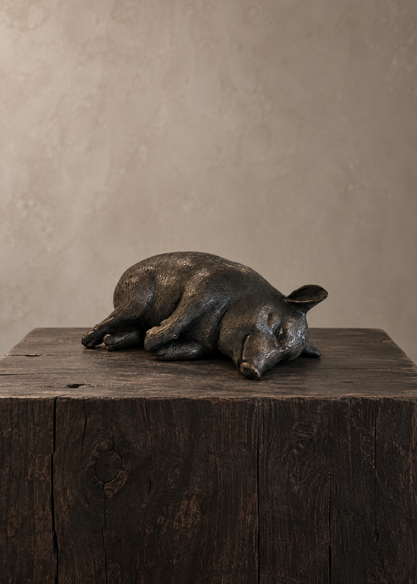 A dark bronze sculpture of a sleeping pig resting on a rustic dark wood block, set against a textured beige background.