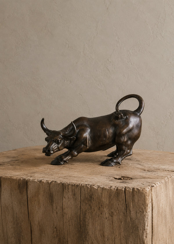 A dark bronze bull sculpture in a crouching, charging pose, displayed on a rustic wooden block against a textured beige wall.
