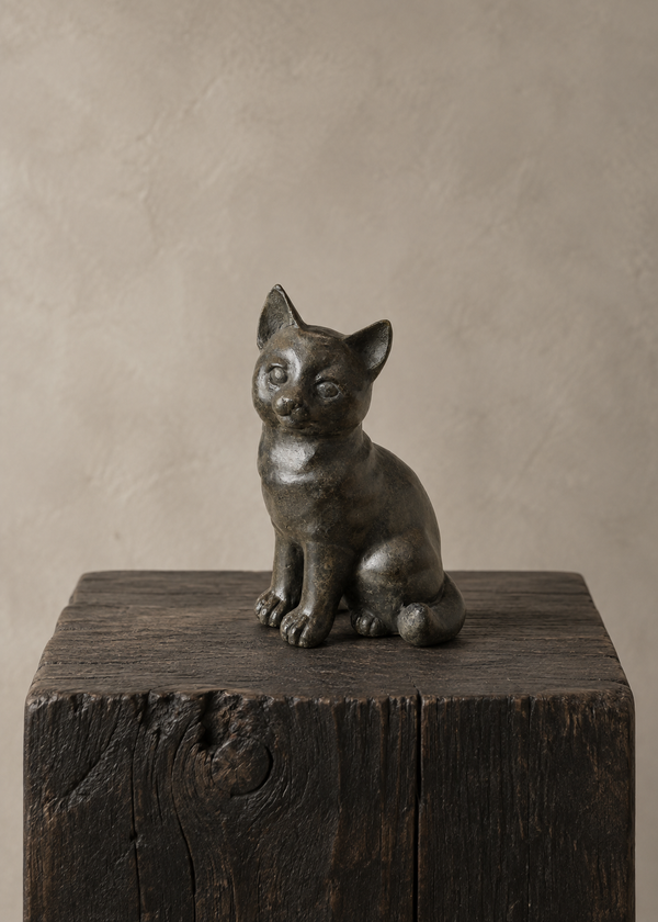 A small dark bronze cat figurine or sculpture sitting on a rustic dark wooden block against a warm beige textured studio background.