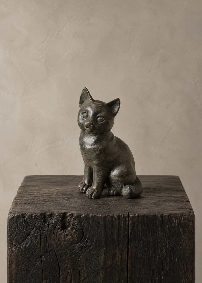 A small dark bronze cat figurine or sculpture sitting on a rustic dark wooden block against a warm beige textured studio background.