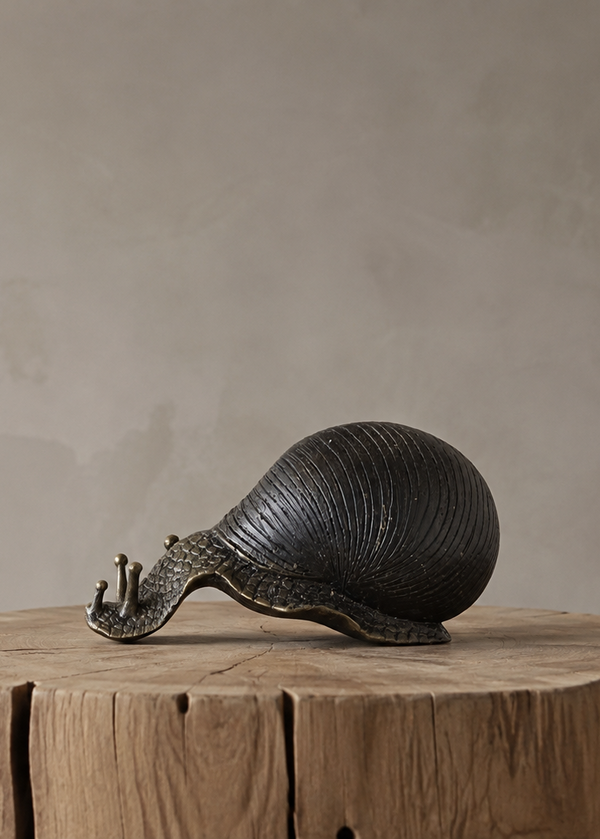 A dark bronze snail sculpture displayed on a rustic wooden stump pedestal against a textured gray concrete wall.