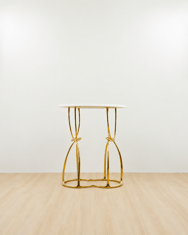 A modern console table with a white oval top and a polished gold brass base featuring twisted, knot-like legs, set against a minimalist white wall and light wood floor.