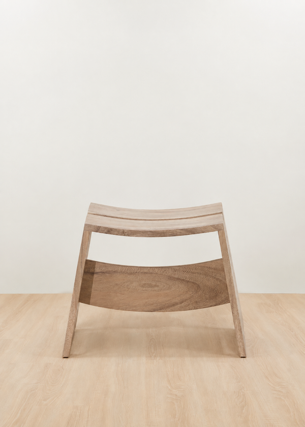 A modern wooden stool with a curved slatted seat and a curved lower support rail, shown in a minimalist studio setting with a light beige wall and pale wood floor.