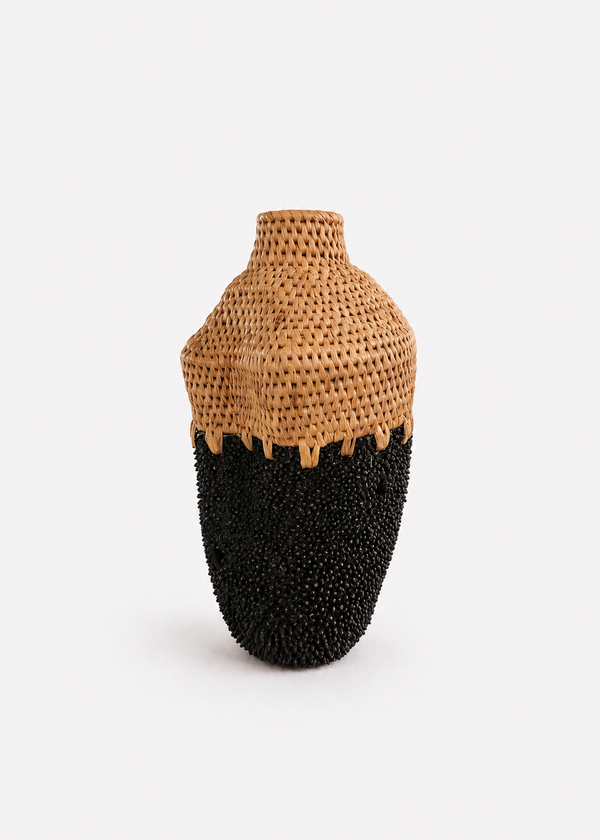 A sculptural ceramic vase with a textured, woven natural tan upper section and a glossy brass black lower section, set against a plain white background.