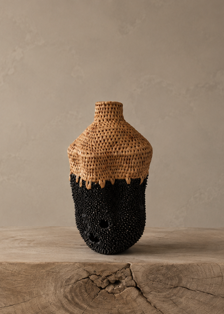 A hand-woven basket vase with a natural tan woven top and a black brass spiky textured bottom, photographed on a wood pedestal and neutral background.