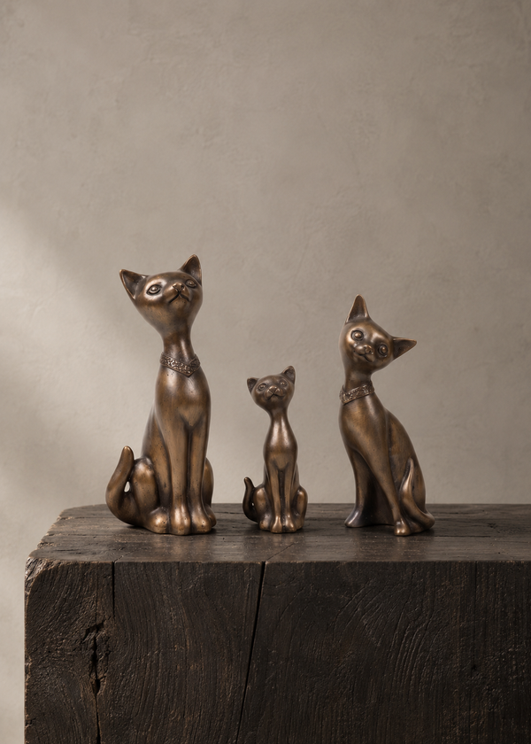Three bronze cat figurines of different sizes arranged on a dark wooden block against a textured beige wall.