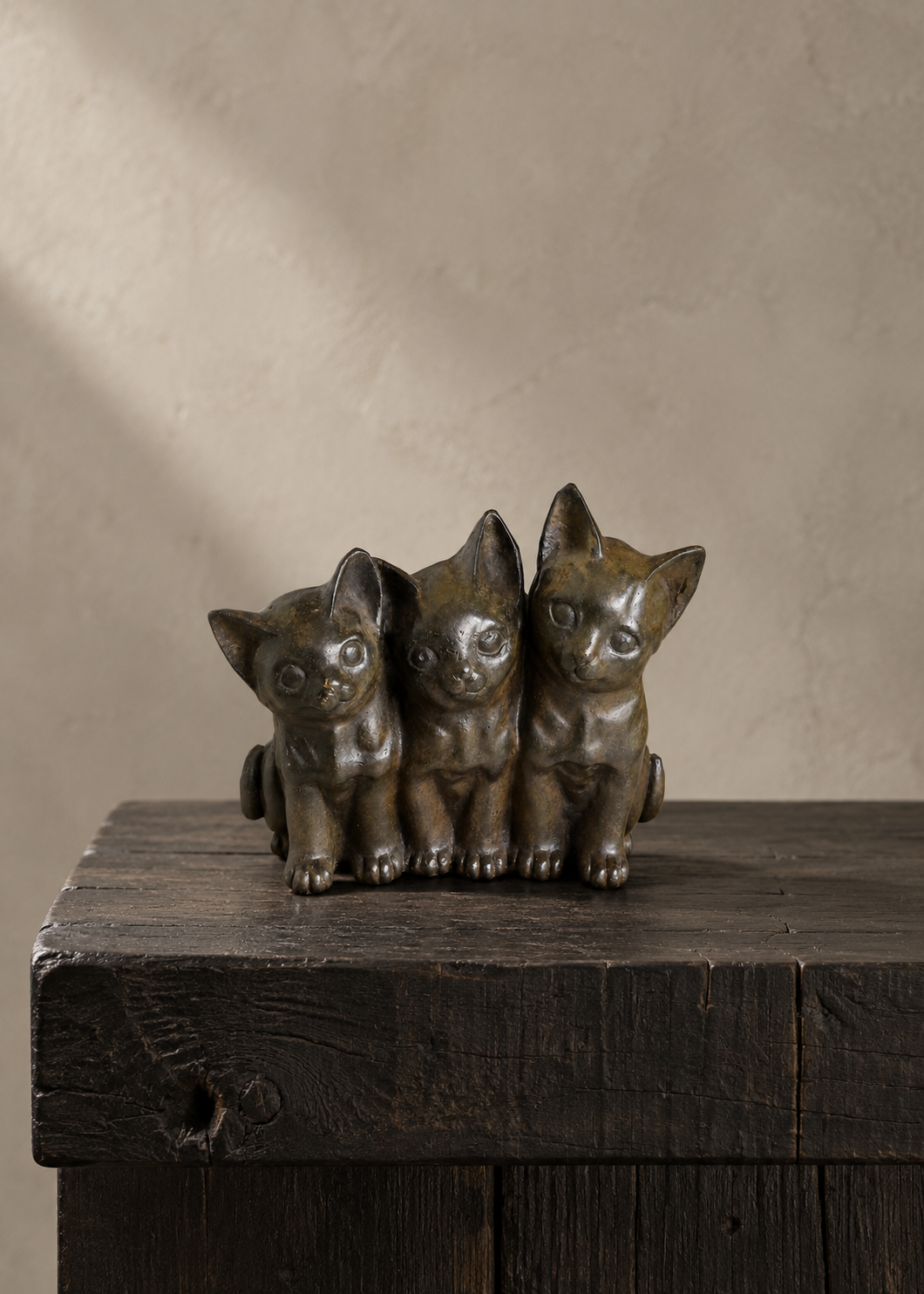Three small bronze kitten figurines sitting side by side on a rustic dark wooden surface against a textured beige wall.