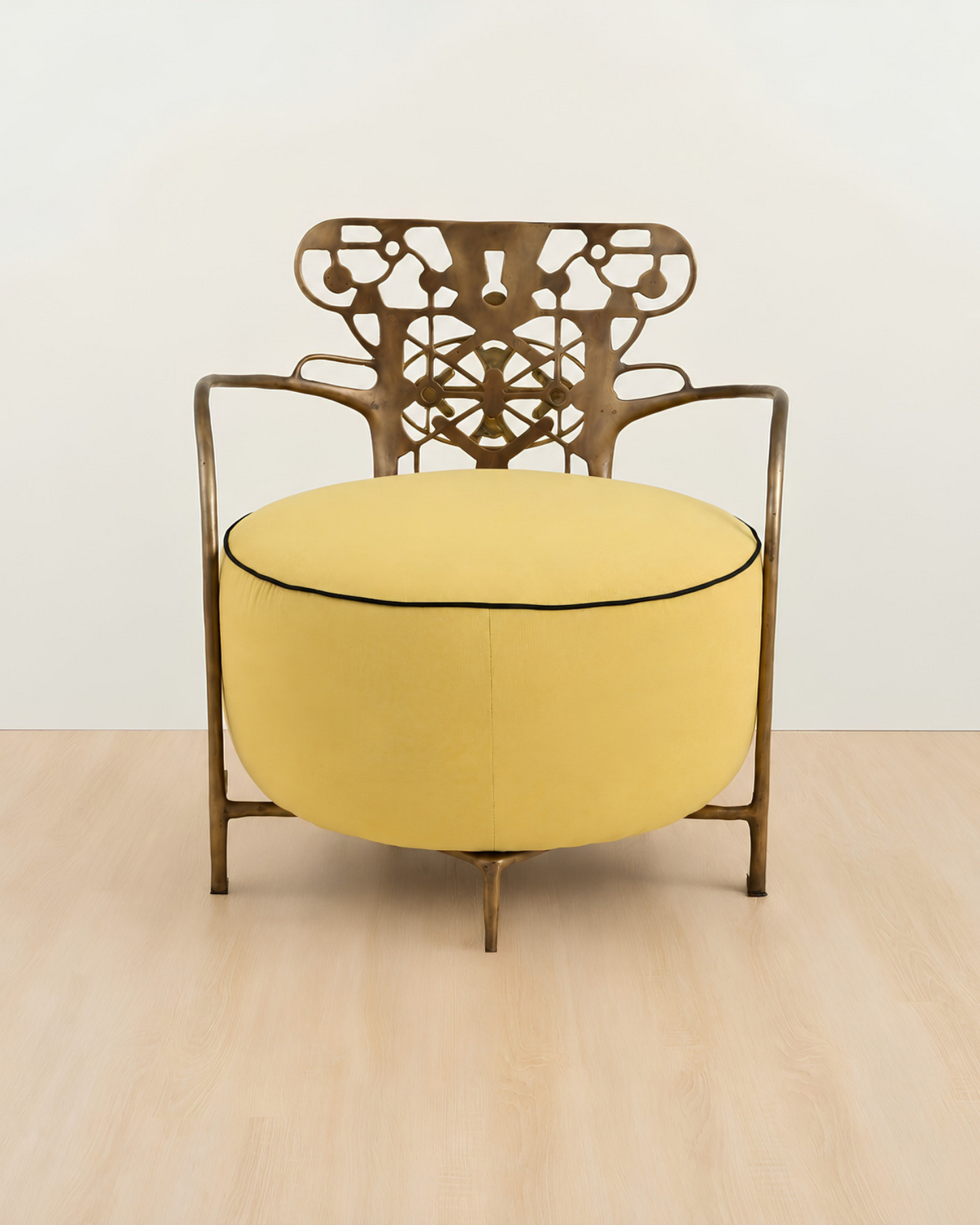 A modern accent chair with a round yellow upholstered seat cushion and a bronze-finished metal frame featuring an intricate cutout backrest, set against a minimalist off-white wall and light wood floor.