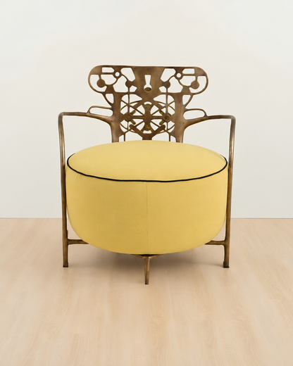 A modern accent chair with a round yellow upholstered seat cushion and a bronze-finished metal frame featuring an intricate cutout backrest, set against a minimalist off-white wall and light wood floor.
