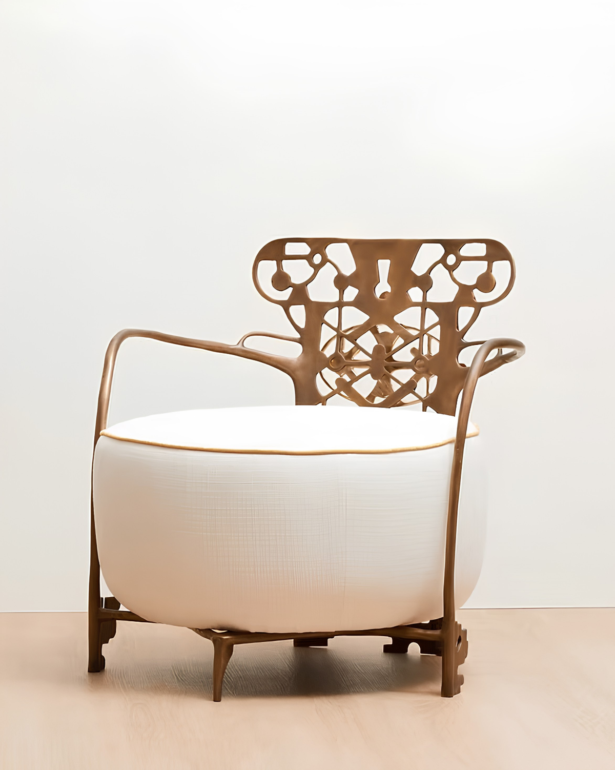 A modern lounge chair with a large white cylindrical cushion, a sculptural bronze metal frame, and a decorative cutout backrest, set against a plain white wall and light wood floor.