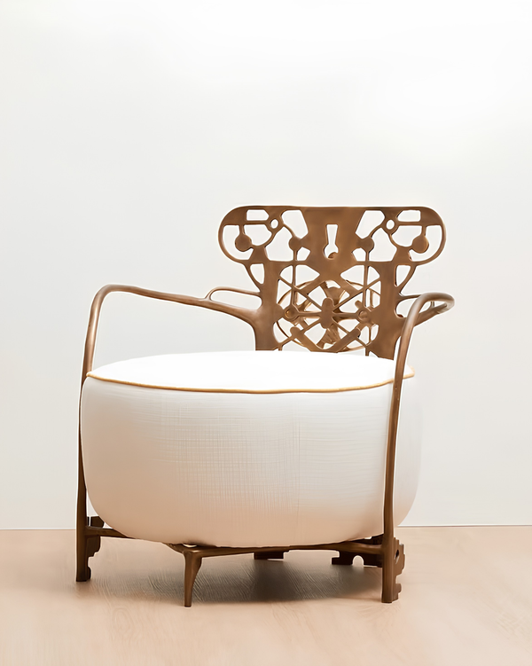 A modern lounge chair with a large white cylindrical cushion, a sculptural bronze metal frame, and a decorative cutout backrest, set against a plain white wall and light wood floor.