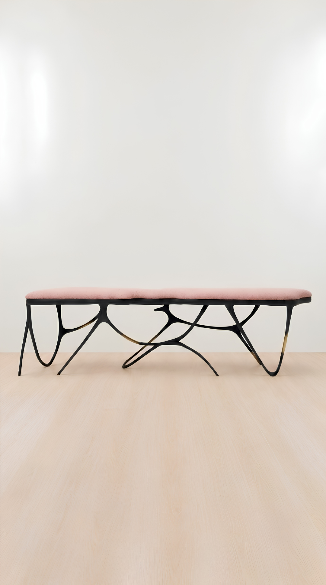 A modern bench with a dusty pink upholstered seat and a sculptural black metal base, shown against a minimalist white wall and light wood floor.