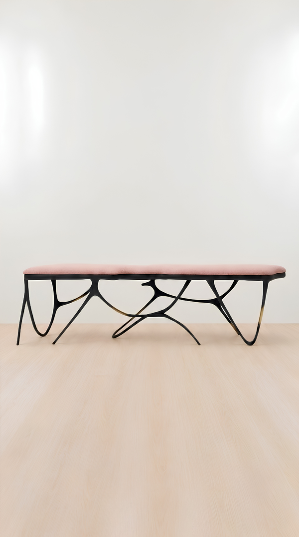 A modern bench with a dusty pink upholstered seat and a sculptural black metal base, shown against a minimalist white wall and light wood floor.