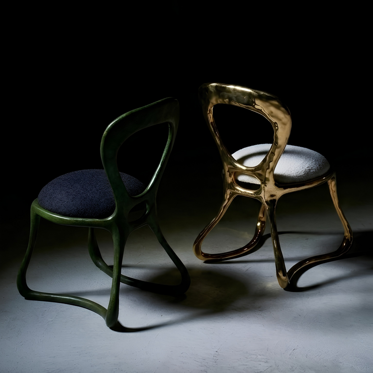 Two sculptural accent chairs displayed on a dark studio floor under dramatic spotlighting. One chair has a dark green frame with a dark blue upholstered seat, and the other has a metallic gold frame with a light gray upholstered seat.