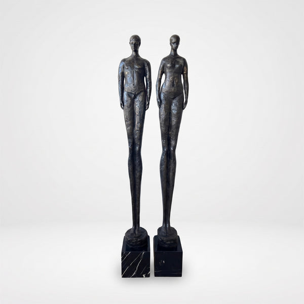 Two dark metallic female figurine sculptures standing side by side on black marble bases against a light gray studio background.