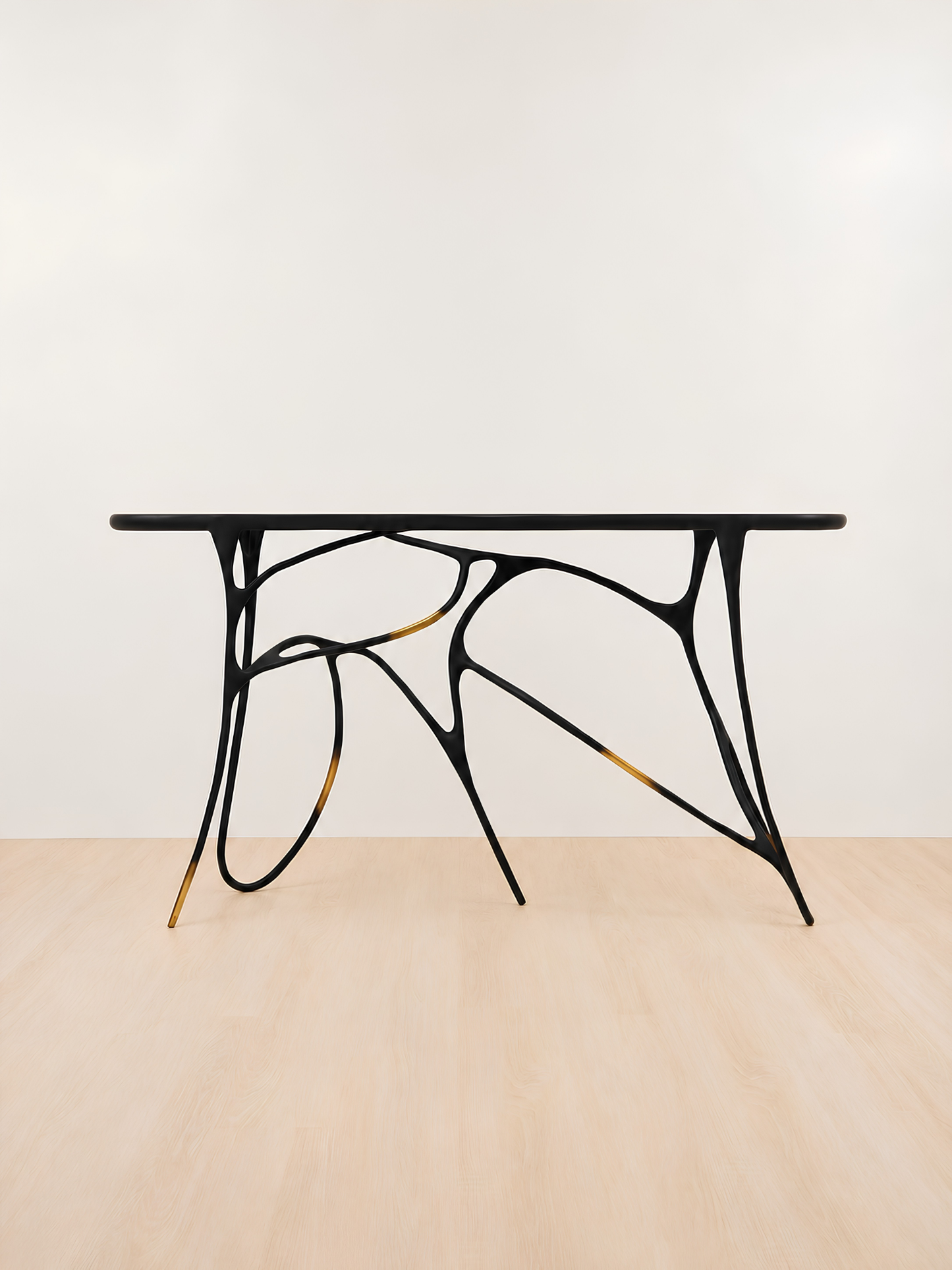 A modern console table with a black rectangular top and an organic, branching black metal base accented with gold-toned details, set against a minimalist off-white wall and light wood floor.