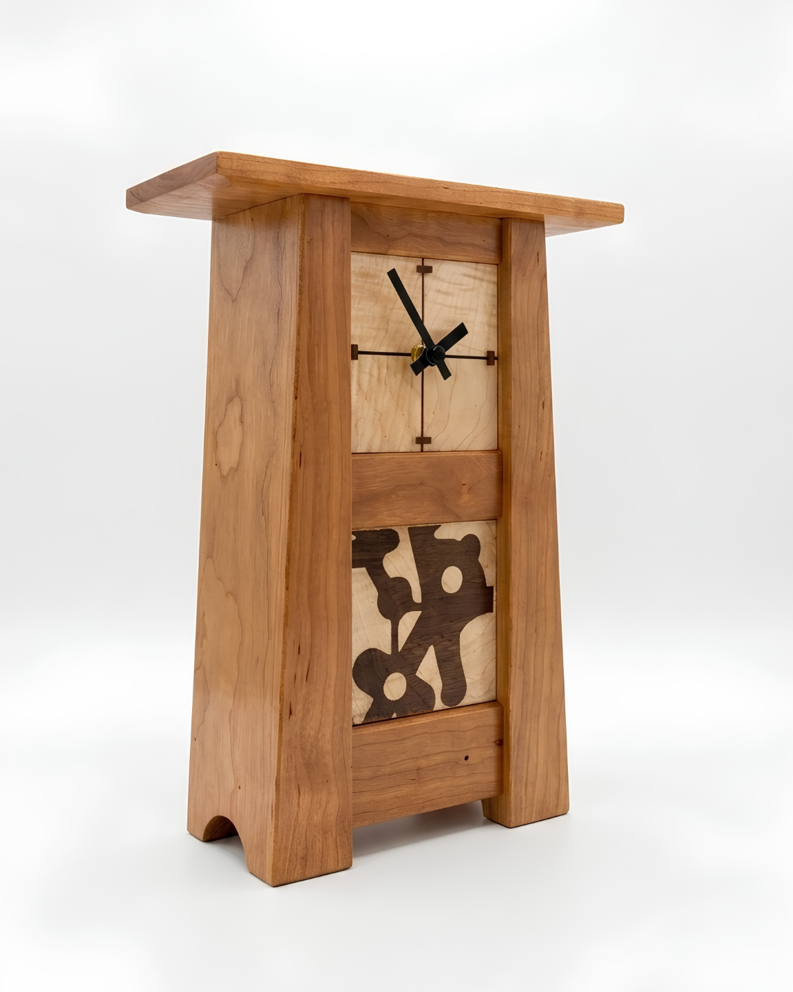 A wooden pedestal clock with a natural oak finish, featuring a central square clock face with black hands and an abstract dark wood inlay on the lower front, set against a plain white background.