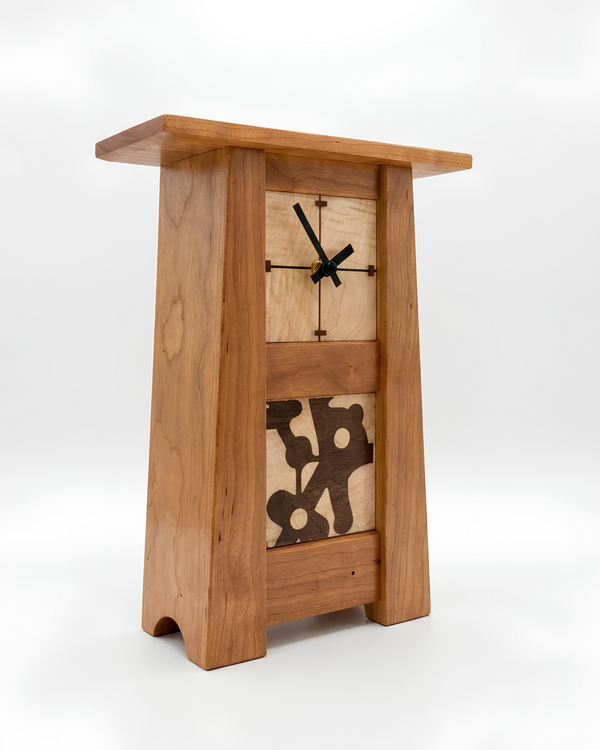 A wooden pedestal clock with a natural oak finish, featuring a central square clock face with black hands and an abstract dark wood inlay on the lower front, set against a plain white background.